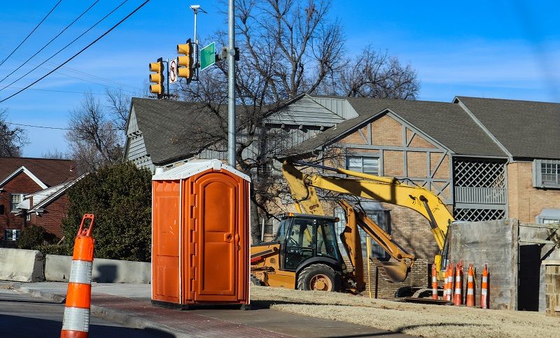 Portable-bathroom-at-construction-site-at-intersectio-of-urban-roads-with-backhoe-in-background-and-traffic-cones-around-cm