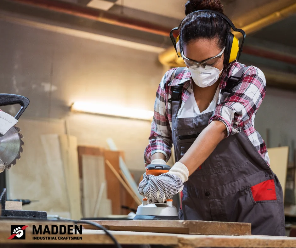 A skilled trades staffing professional sanding wood in a workshop, wearing safety gear including gloves, goggles, and earmuffs